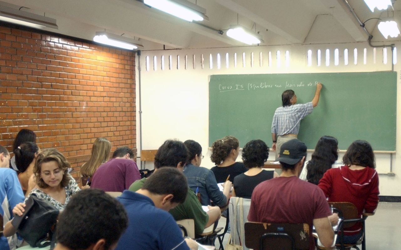 Professor escreve no quadro enquanto estudantes acompanham uma aula, em fotografia de arquivo.